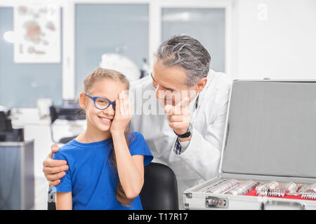 Reifen Augenarzt in weißen Mantel Prüfung des Sehvermögens der hübschen Mädchen in der Medizinischen Klinik. Lächelnd patient Kind sitzen in Blau Brille und schließen ein Auge mit der Hand. Stockfoto