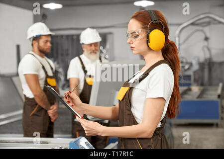 Ingenieurin im schützenden Gehörschützer und Gläser Ordner, schreiben. Ingenieure tragen in den weißen T-Shirts, Overalls, Helme. Betrieb edv-Plasma laser Maschine. Stockfoto