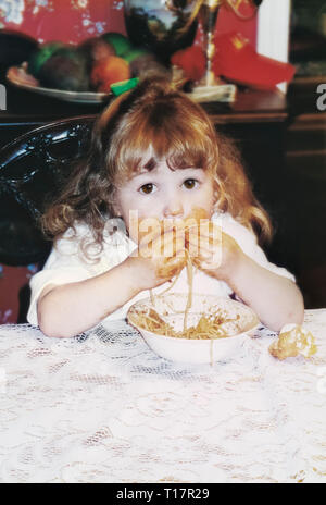 Niedlich lustig chaotisch Kleinkind Mädchen Alter 2 mit langen Lockigen, blonden Haaren und großen ausdrucksvollen braunen Augen Essen Nudeln und Spaghetti alle über ihr Gesicht Stockfoto