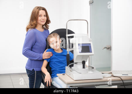 Hübsches Mädchen im Stuhl sitzend am Tisch mit modernen Spaltlampe Maschine für die Prüfung des Sehvermögens. Mutter und Kind posiert in Schöne neue Optiker Büro der Augenheilkunde Klinik. Stockfoto