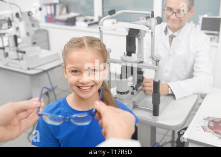 Augenarzt, blaue Brille zu Patienten. Hübsches Mädchen lächelnd und Suchen an der Kamera, in der Klinik darstellen. Arzt Augenarzt am Tisch sitzen mit spaltlampe auf Hintergrund in der Klinik. Stockfoto