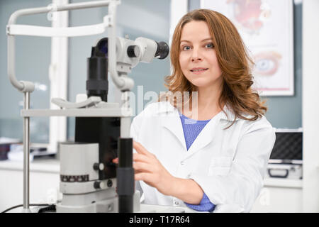 Schöne Frau, Augenarzt sitzen, an der Kamera suchen, die mit Medical optometry Ausrüstung der für die Prüfung des Sehvermögens posieren. Professionelle Augenarzt in medizinischen Zimmer der Klinik. Stockfoto