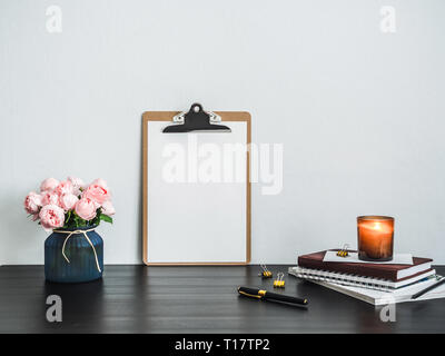 Zwischenablage mit weiße Leere Seite auf dem Tisch. Zwischenablage, rosa Blumen, duftende Kerze, Stapel von Büchern auf Schwarz tabetop. Zwischenablage mock up mit leeren Blatt Papier A4 Hochformat, kopieren Platz für Text Stockfoto