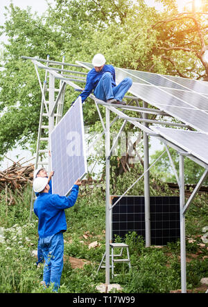 Installieren von stand-alone-Solar Photovoltaik Panel System. Arbeitnehmer in Hard - Hüte und Blau insgesamt heben das Solarmodul auf Metall-Plattform. Alternativ Stockfoto