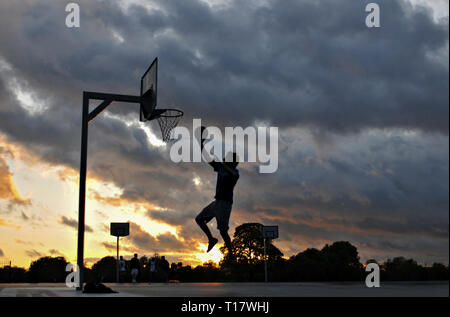 Ein Mann, gegen eine stürmische Sonnenuntergang Silhouette, spielt Basketball außerhalb Stockfoto