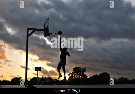 Ein Mann, gegen eine stürmische Sonnenuntergang Silhouette, spielt Basketball außerhalb Stockfoto