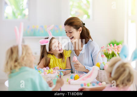 Mutter und Kinder Farbe Ostereier. Mama, kleine Mädchen und Jungen mit Hasenohren sterben und Malerei für die Ostereiersuche im Weißen sonnige Zimmer. Familie Feier Stockfoto