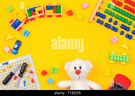 Kinder Spielzeug Hintergrund. Teddybär, Holzeisenbahn, bunte Bausteine, Spielzeug tools Kit, Autos, Abacus auf gelben Hintergrund. Ansicht von oben Stockfoto