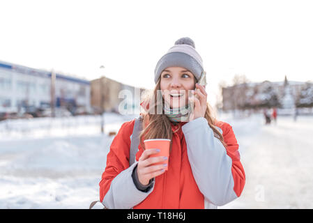 Mädchen überrascht Winter in der Stadt an der frischen Luft auf dem Hintergrund von Schnee und Schneeverwehungen, fordert das Telefon, freudig und fröhlich durch den Anruf schockiert, in Ihrem Stockfoto