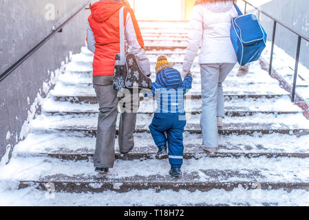 Eltern Mutter und Freundin Mädchen halten Hand kleiner Junge Sohn 3-6 Jahre alt, und gehen Sie die Treppe im Übergang, winter Stadt Hintergrund Schnee Matsch. Ansicht von der Rückseite, in Stockfoto