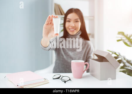 Schönes Bild von attraktiven Mädchen sitzen am Tisch und zeigen die Schönheit Produkt auf die Kamera. Es ist die Hand Creme. Junge Frau lächelt und schaut Stockfoto