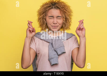 Lustige und niedliche Mädchen kreuzt die Finger an beiden Händen und halten ihre Augen geschlossen. Sie hofft. Auf gelbem Hintergrund Stockfoto