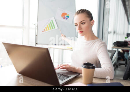 Mädchen ist auf der Suche nach Laptop und demonstrieren Diagramme auf den Schreibtisch. Sie arbeitet jetzt. Junge Frau blickt sehr ernst Stockfoto