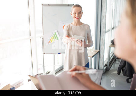 Fröhlich und glücklich Lautsprecher ist in der Nähe von standin Flipchart mit Diagrammen. Sie wartet, während das Mädchen mit Notebook auf Ihre Frage beantworten wird. Stockfoto