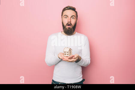 Erstaunt Kerl steht und mit einem Holz Spielzeug Haus. Er sucht gerade nach vorn. Auf rosa Hintergrund isoliert Stockfoto