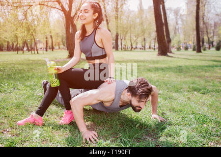 Lustiges Bild junger Mann und Frau. Er tut Push-ups und gerade auf der Suche, während Sie auf ihm sitzt und lacht. Auch Mädchen hält einen bottl Stockfoto