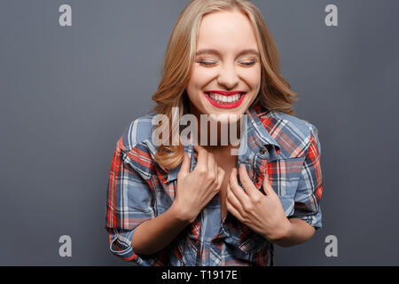 Nette und hübsche junge Frau lacht. Sie hat ihre Augen geschlossen. Mädchen hält ihre Hände in der Nähe der Brust. Auf grauem Hintergrund Stockfoto