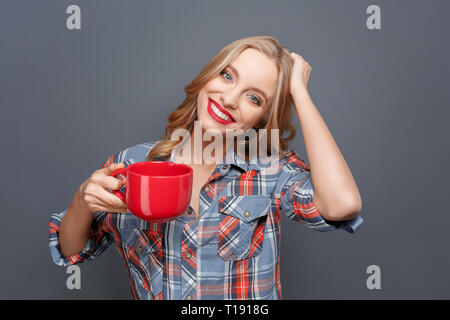 Junge und hübsche Frau hält einen roten Becher in der rechten Hand und hält die andere Hand auf das Haar. Sie sieht glücklich aus. Auf grauem Hintergrund Stockfoto