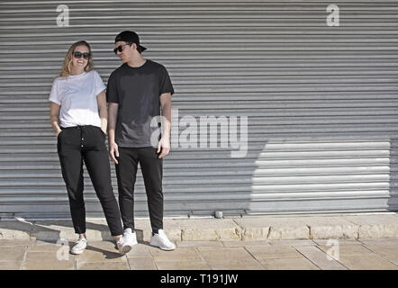 Junge Natürliche und Ungezwungene sportliche Paar gekleideter Mann mit Kappe nach hinten und Frau gegen Wellblech wall street style Stockfoto