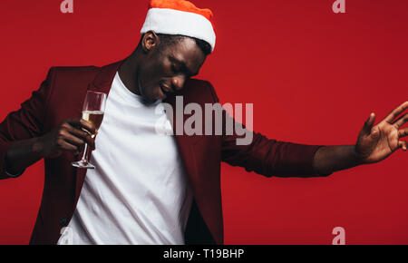 Afrikanischen Mann, santa hut mit Champagner tanzen gegen roten Hintergrund. Glückliche junge schwarze Kerl genießt bei der Weihnachtsfeier. Stockfoto