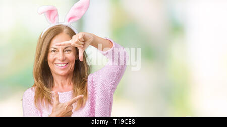 Mittleres Alter brünette Frau bekleidet Osterhasen Ohren über isolierte Hintergrund lächelnd, Rahmen mit Händen und Fingern, glückliches Gesicht. Kreativität Stockfoto