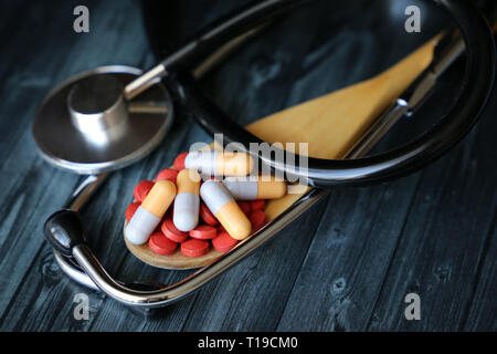 Pillen und Kapseln in Löffel aus Holz und Stethoskop auf dunklen Holztisch. Konzept der Gesundheit, Drogen, Blutdruck, Vitamine, Apotheke, Behandlung Stockfoto