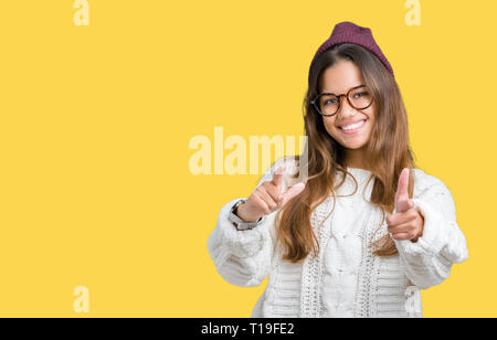 Junge schöne Brünette hipster Frau Brille trägt und im Winter hat über isolierte Hintergrund Pointing Finger Kamera mit glücklich und lustiges Gesicht. G Stockfoto