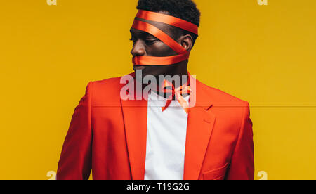 Afrikanischer Mann mit Satinband auf seinem Gesicht gegen den gelben Hintergrund gewickelt. Mann in orange Blazer verpackt als Geschenk. Stockfoto
