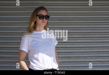 Junge casual sportlich gekleidet blonde Frau mit Sonnenbrille im weißen T-Shirt gegen Wellblech wall street style Stockfoto