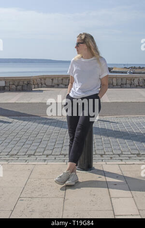 Junge casual sportlich gekleidet blonde Frau mit Sonnenbrille im weißen T-Shirt sitzt in der Nähe der Straße mit Blick auf das Meer hinter auf Mallorca, Spanien. Stockfoto