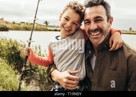 Porträt ein Mann stand am Ufer eines Sees mit seinem Kind eine Angelrute. Lächelnd Vater und Sohn in der Nähe von einem See miteinander halten. Stockfoto