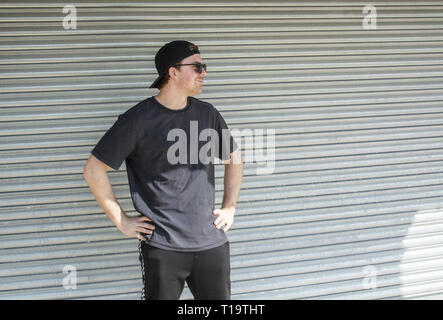 Junge casual Sportlich gekleideter Mann mit Kappe nach hinten in Schwarz gegen Wellblech wall street style Stockfoto