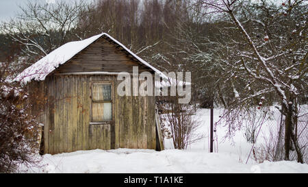 Ein kleines altes Holzhaus mit einem Fenster, ein Zaun und ein kleiner Garten im Winter mit Schnee bedeckt Stockfoto