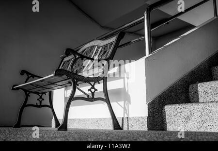 Leere Schmiedeeisen Sitzbank in der Nähe der Treppe des Gebäudes platziert. Sonnenschein in der konkreten Gebäude. Deprimiert, traurig und einsam. Eine leere wrough Stockfoto