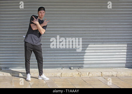 Junge casual Sportlich gekleideter Mann mit Kappe nach hinten in Schwarz gegen Wellblech wall street style Stockfoto