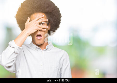 Junge afrikanische amerikanische Mann mit afro Haar Brille peeking im Schock für Gesicht und Augen mit der Hand, schauen durch die Finger mit verlegen Stockfoto