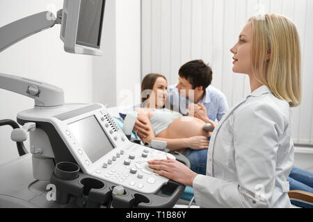 Ultraschall-untersuchung in der modernen medizinischen Klinik. Professionelle Arzt mit Ultraschall, bei Monitor suchen. Glückliches Paar. Schwangere Frau liegend, Mann die Hand der Frau. Stockfoto
