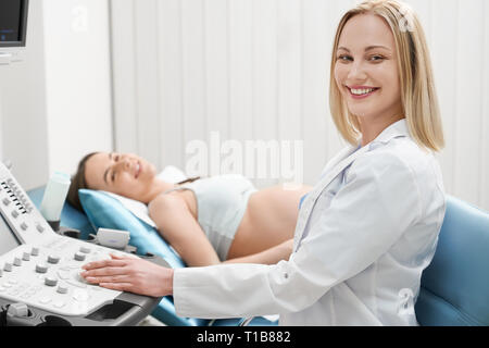Schöne, hübsche Blondine Arzt tragen in Weiß Medical Gown, Ultraschall Untersuchung der Frau Bauch, bei Camera suchen, lächelnd. Schwangere Frau liegen. Konzept des modernen, professionellen Klinik. Stockfoto