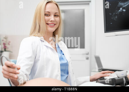Charmant, schöne blonde Frau an der Kamera suchen, lächeln und posieren. Ärztin tragen in Weiß Medical Gown Haltevorrichtung, Ultraschalluntersuchung in der modernen Klinik. Stockfoto