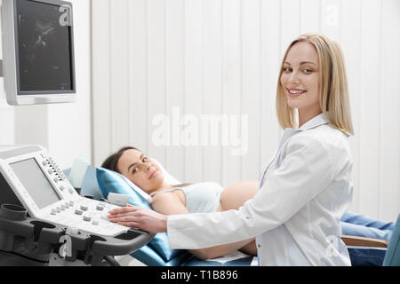 Schöne, hübsche Blondine Arzt tragen in Weiß Medical Gown, Ultraschall Untersuchung der Frau Bauch. Schwangere Frau liegend, mit Blick auf die Kamera lächelnd. Modernes Ultraschallgerät. Stockfoto