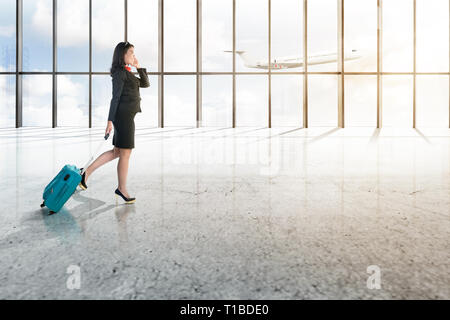 Junge asiatische Geschäftsmann mit Mobiltelefon und blauen Koffer auf dem Flughafen Halle mit Fenster aus Glas und fliegende Flugzeug über blauen Himmel backgro Stockfoto