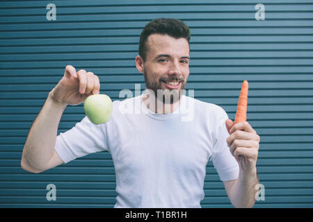 Glücklich Mann steht und Halten von Obst und Gemüse in den Händen. Es ist grüner Apfel und Orange Karotte. Er ist auf der Suche auf Kamera und lächelnd. Kerl sieht Stockfoto