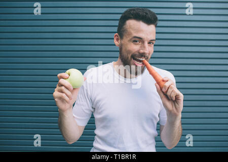 Emotionale Mann im weißen Hemd steht und auf der Suche nach Kameras. Er lächelt und posieren. Kerl, biss Karotte und Holding grüner Apfel in der anderen Hand Stockfoto