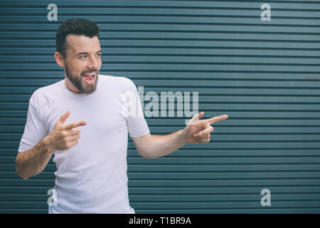 Schön und positiver Mann lächelt und poitning mit Fingern auf der rechten Seite. Bärtiger Kerl schaut aufgeregt. Auf gestreifte und blauem Hintergrund isoliert. Stockfoto