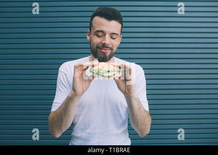 Bärtiger Mann hält Burger mit beiden Händen. Er sucht ihn an und lächelte. Auf gestreifte und blauem Hintergrund isoliert. Stockfoto