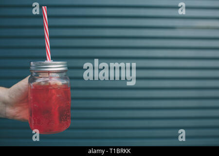 Ein Bild von der Hand des Menschen halten Glas Tasse rote Limonade. Es hat Stroh. Auf gestreifte und blauem Hintergrund isoliert. Stockfoto