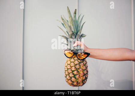 Ein Bild von Ananas mit Sonnenbrille auf. Woman's Händen hält. Auf gestreift und weißem Hintergrund. Stockfoto