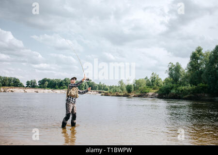 Ein Bild von Mann in Wasser und winkte mit der Fliegenrute. Er hält mit beiden Händen. Kerl sucht zur Rute. Er trägt eine Sonnenbrille, Weste und Watvögel. Stockfoto