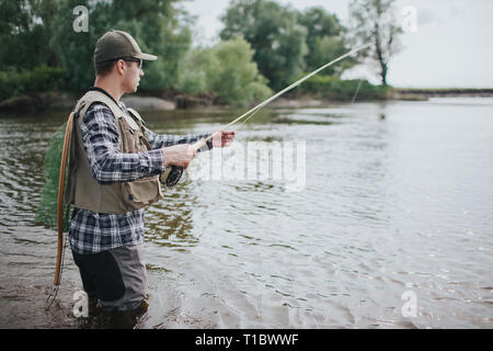 Ernster Mann steht in Wasser und Angeln. Er hat Spinnen in die Hände und Angeln net auf der Rückseite. Kerl trägt Weste, Watvögel, T-Shirt und Cap. Stockfoto