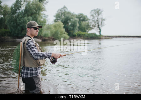 Fischer in Sonnenbrille steht in Wasser. Er schaut nach rechts. Kerl hat die Fischerei net auf der Rückseite. Der Mensch ist die Rute mit einer Hand. Er sieht Stockfoto